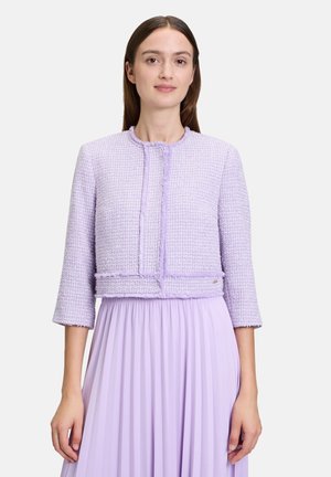 Veste en tweed lavande avec des manches trois-quarts, tissu texturé, design sans col et garniture frontale. Portée sur une jupe plissée lavande.