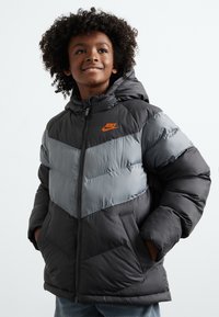 Puffer takki, jossa on musta pohjaväri ja harmaat viistopaneelit. Takissa on huppu, vetskari ja rinnassa oranssi Nike-logo.