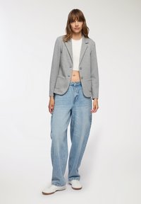 Blazer grigio su misura con due bottoni e tasche laterali, abbinato a jeans azzurri larghi e sneakers bianche.