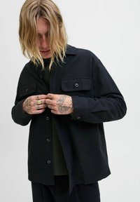 Schwarzes Hemd mit Knopfleiste, entspannter Passform, zwei Vordertaschen und weicher Textur, getragen über einem grünen Pullover.