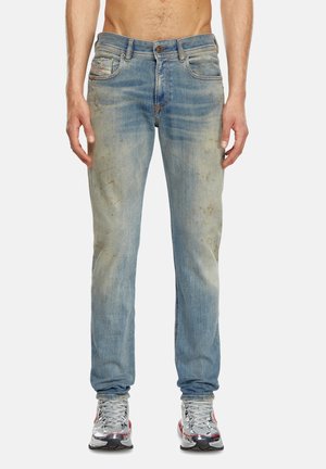 Homme portant un jean bleu délavé, légèrement taché, et des chaussures de course argentées, debout devant un fond clair uni.