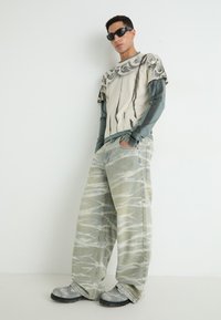 Overdimensjonert beige og grå mønstret T-skjorte lagd over en langermet teal skjorte, kombinert med grå mønstret vide jeans og teksturerte sko.