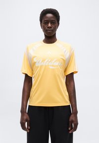 Άτομο με κοντά μαλλιά που φοράει κίτρινο αθλητικό πουκάμισο Adidas και μαύρο παντελόνι, στέκεται μπροστά σε απλό φωτεινό φόντο.