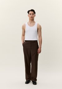 Mlad moški stoji, oblečen v belo majico brez rokavov, rjave hlače in črne čevlje, na enostavni svetli podlagi.