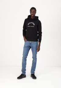 Sort hættejakke med hvidt logo, blå jeans og sorte sneakers. Modellen står i en neutral position på en ensfarvet baggrund.