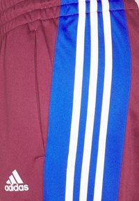 Pantalon de sport Adidas bordeaux avec des panneaux bleus présentant trois bandes blanches verticales et un logo Adidas blanc près de la poche.