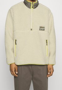 Sudadera de felpa color crema con cuello gris, acentos en amarillo neón, bolsillos con cremallera y un parche en el pecho con un diseño gráfico.