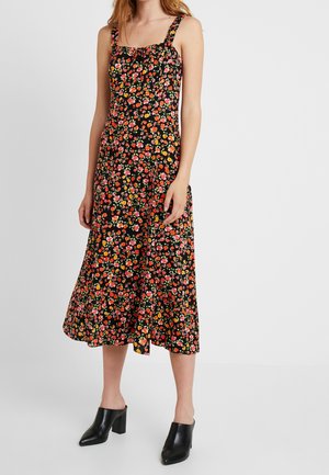 Vrouw draagt een mouwloze zwarte midijurk met rode, oranje en roze bloemenprint en zwarte mules met hak.