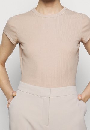 Osoba w beżowej, prążkowanej koszulce z krótkim rękawem i jasnobeżowych spodniach z wysokim stanem, z rękami w kieszeniach, na białym tle.