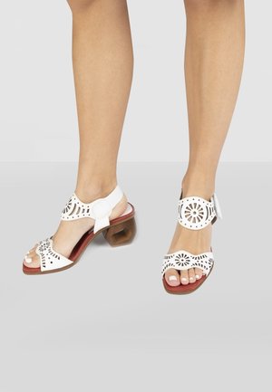 Bílé sandály mají výřezové vzory, zadní pásek a šestiúhelníkový dřevěný podpatek. Podešev je červená a prsty jsou odkryté.