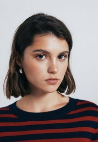 Model met kort bruin haar, draagt een gestreepte trui in rood en marineblauw. Heeft grote zilveren creolen en een natuurlijke maquillage.