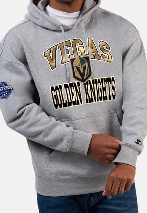 Mężczyzna ubrany w szarą bluzę z kapturem z napisem i logo „Vegas Golden Knights”, naszywką Konferencji Zachodniej NHL na rękawie, poprawiający mankiet.