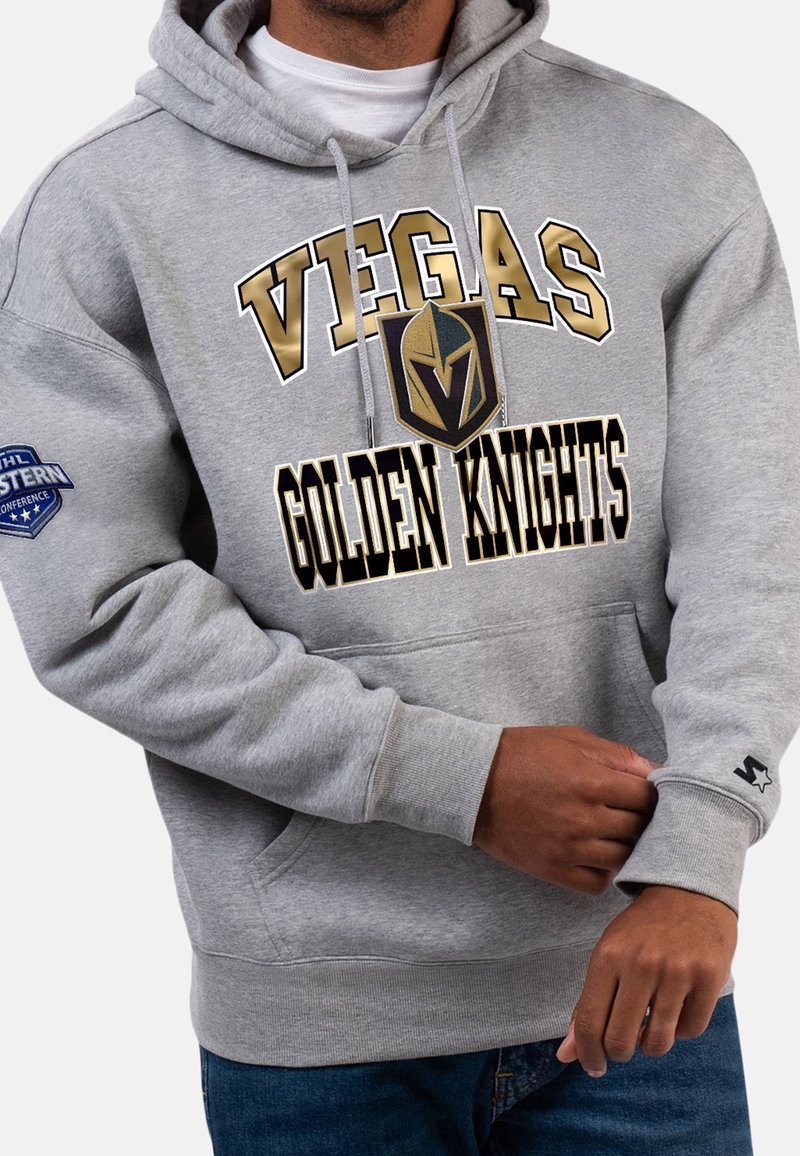 Homme portant un sweat à capuche gris avec l'inscription "Vegas Golden Knights" et le logo, patch de la Conférence Ouest de la NHL sur la manche, ajustant le poignet.