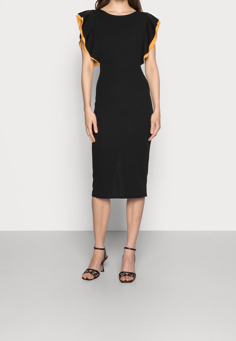 Femme portant une robe noire mi-longue avec des manches à volants orange et des sandales noires à talons hauts à lanières, debout devant un fond uni.