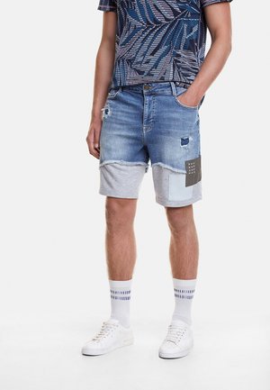 Homme portant un short en denim bleu et en patchwork gris avec des déchirures, des chaussettes blanches à rayures bleues, des baskets blanches et un t-shirt bleu à motifs.