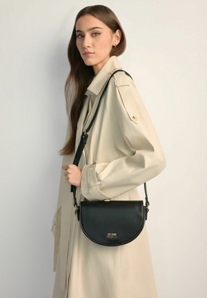 Kobieta o długich brązowych włosach, ubrana w beżowy płaszcz trencz, niosąca czarną torebkę na ramię w kształcie półksiężyca z złotym logo z przodu.