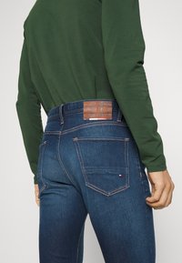 Tmavě modré džíny z denimu s hnědou koženou nášivkou, vybavené zadními kapsami a kontrastním prošitím. Materiál vypadá hladký a odolný.