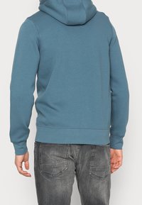 Sudadera con capucha en un tejido azul verdoso, con una textura suave y dobladillo acanalado. Combinada con unos jeans gris oscuro con bolsillos traseros.