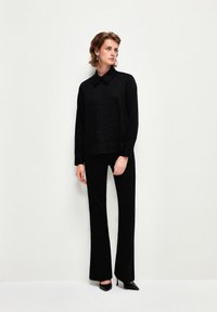 Camicia nera con colletto e una sottile texture abbinata a pantaloni neri svasati e scarpe con punta appuntita. Design minimalista, monocromatico.