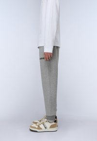 Graue Sweatpants mit elastischen Bündchen, die mit einem Logo-Patch am Oberschenkel versehen sind, kombiniert mit beigen und weißen Sneakern. Ein langärmliges, weißes Oberteil ist sichtbar.