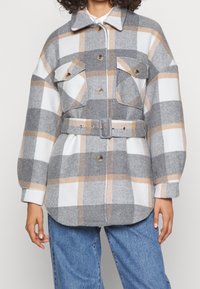 Chaqueta de cuadros gris y blanco hecha de una mezcla de lana, con dos bolsillos en el pecho, una cintura ceñida con cinturón y un collar desplegado.