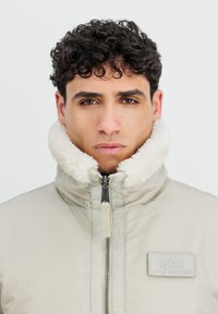 Bežna jakna na zadrgo z visokim, puhastim belim ovratnikom in teksturirano nalepko z oznako "Alpha Industries" na prsih.