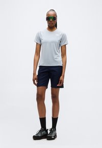 Γυναίκα που στέκεται, φοράει κοντομάνικο γκρι ανοιχτό χρώμα, σκούρα μπλε σορτς, μαύρες ψηλές κάλτσες, γκρι παπούτσια πεζοπορίας και μεγάλα αντανακλαστικά αθλητικά γυαλιά ηλίου.