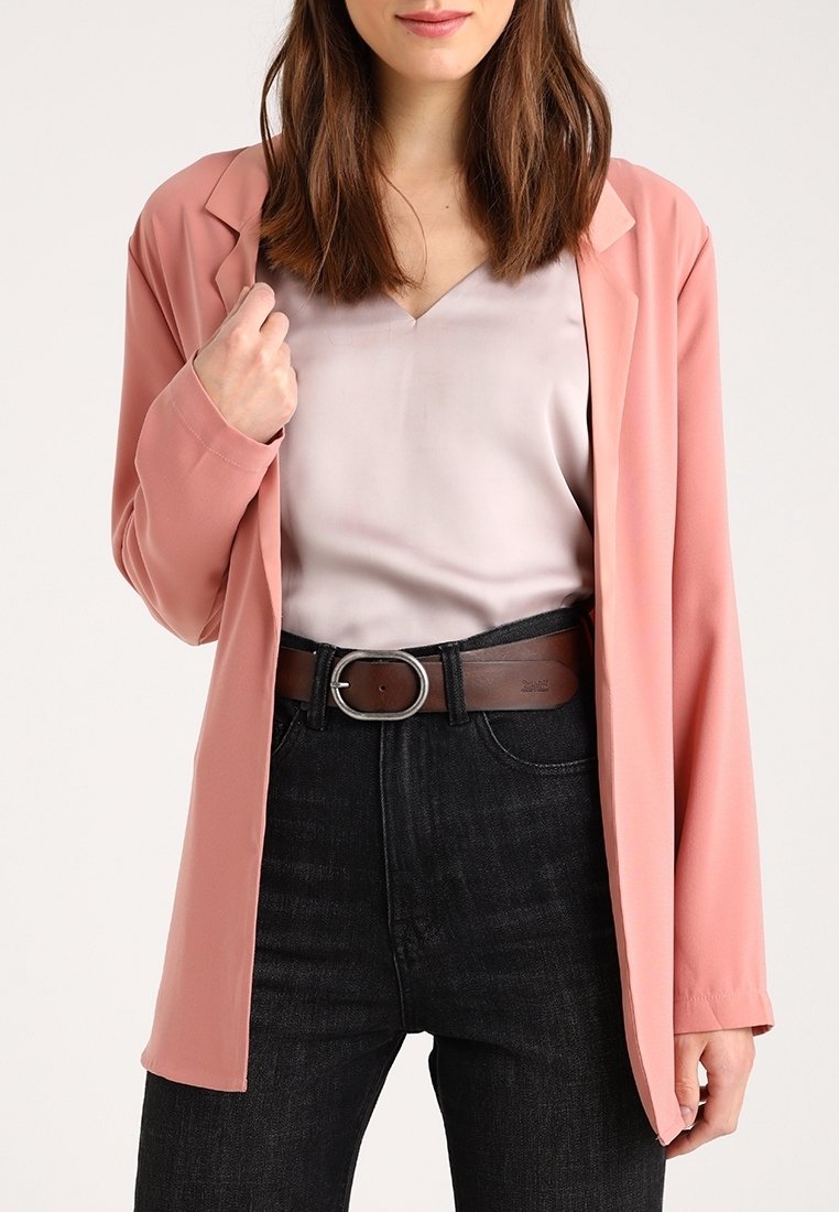 Femme portant un blazer rose clair, un chemisier lavande pâle, un jean taille haute foncé et une ceinture marron avec une boucle argentée.