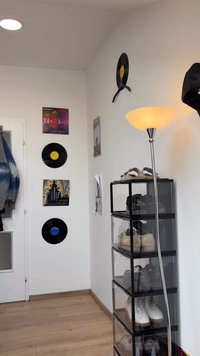 Una lampada da terra cromata con un paralume bianco si trova accanto a un'unità di stoccaggio nera. La parete è decorata con dischi in vinile e vari poster.