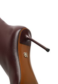 Braune Lederschuhe mit einem geschwungenen Design, das ein goldfarbenes Logo-Element und einen schwarzen Absatz aufweist. Glänzende Oberfläche und kontrastierende Farbbereiche.