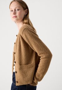 Donna con capelli chiari che indossa un cardigan marrone strutturato con bottoni sopra una maglietta bianca e jeans blu scuro, su uno sfondo chiaro e uniforme.