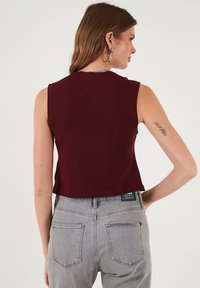 Bordový bezrúkový crop top vyrobený z jemného materiálu, s okrúhlym výstrihom a čistým lemovaním, v kombinácii so svetlo šedými vysokými džínami.