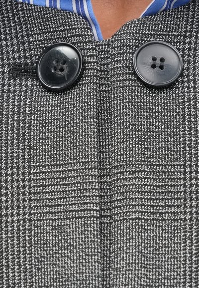 Grijs houndstooth-patroon stof heeft twee zwarte knopen bij de halslijn. Een blauw-wit gestreept shirt is eronder zichtbaar.