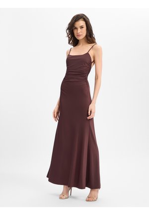 Frau trägt ein ärmelloses, bodenlanges dunkelbraunes Kleid mit dünnen Trägern und gerafften Seiten, steht mit einer Hand locker seitlich.