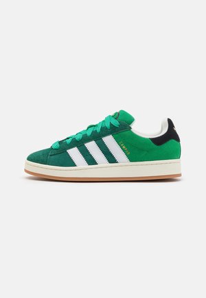 Zielony zamszowy sneaker Adidas Campus z białymi paskami, zielonymi sznurówkami, gumową podeszwą i czarną zawieszką na pięcie na białym tle.