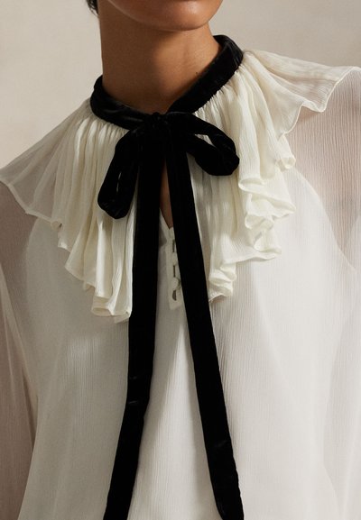 Balta blūze ar melnu samta apkakli, tai ir krokojums priekšpusē un auklas. Audums ir caurspīdīgs ar teksturētu rakstu un pogu detaļām.