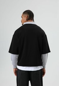 Zwart oversized T-shirt met korte mouwen, gedragen over een grijze lange mouwen shirt. Heeft een brede hals en een losse pasvorm.