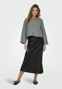 Grauer, gekürzter Pullover mit weiten Ärmeln, kombiniert mit einem schwarzen Satin-Midirock und schwarzen flachen Schuhen mit dekorativen Akzenten.