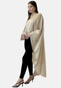 Beżowy satynowy ponczo o luźnym kroju, z szerokimi rękawami i krawędzią w stylu high-low, w zestawieniu z czarnymi legginsami i sandałami na obcasie.