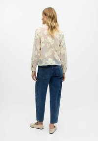 Gennemsigtig, lyse beige bluse med blomsterbroderi, parret med mørkeblå denimbukser. Skoene er lyse med en struktureret finish.