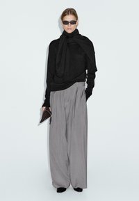 Maglione nero con dettagli al collo annodati, abbinato a pantaloni grigi larghi plissettati. Gli accessori includono occhiali da sole neri e una pochette marrone.