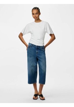 Jeune femme aux cheveux tressés, debout les mains sur les hanches, portant un t-shirt blanc à manches courtes, un jean bleu large et court, et des sandales noires.
