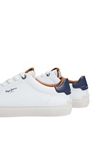 Zapatillas blancas con textura suave, con acentos marrones y azul marino en el talón, y diseño de cordones. Logotipo visible en el lateral.