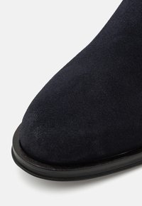 Zwarte suède enkelboot met een ronde neus, gladde textuur en een contrasterende donkere zool, die een minimalistisch ontwerp benadrukt.