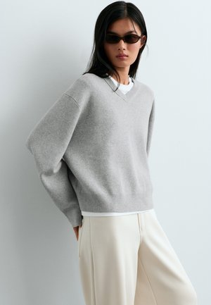 Vrouw met donkere zonnebril, lichtgrijze oversized trui, wit overhemd eronder en offwhite wijde broek tegen een effen lichte achtergrond.