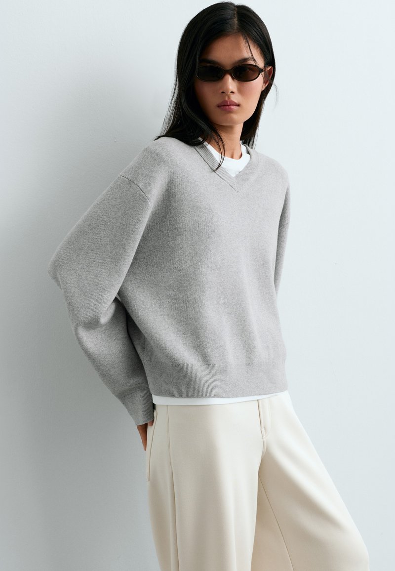 Femme portant des lunettes de soleil foncées, un pull oversize gris clair, une chemise blanche en dessous et un pantalon large écru sur un fond clair uni.