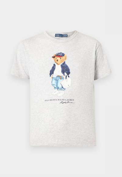 Ανοιχτό γκρι T-shirt με γραφικό ενός αρκούδου που φοράει καπέλο Polo, ναυτικό μπουφάν, τζιν, λευκά αθλητικά παπούτσια και κρατάει λευκή τσάντα.