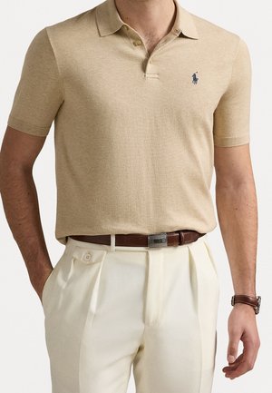 Homme portant un polo beige à manches courtes avec un logo bleu marine, rentré dans un pantalon blanc plissé avec une ceinture en cuir marron et une montre assortie.