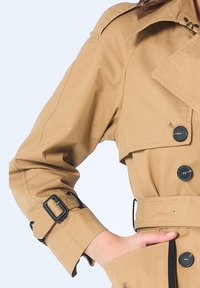 Manteau beige à double boutonnage, grand col et poignets boutonnés. Doté d'une taille ceinturée et d'accents noirs sur les boutons.