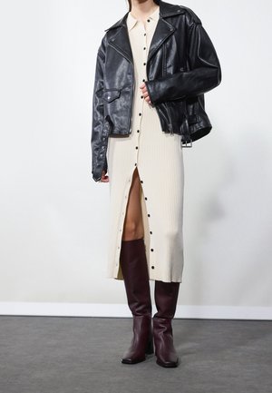 Personne portant une veste en cuir noire sur une robe midi crème à boutons avec une fente frontale, associée à des bottes hautes marron foncé.